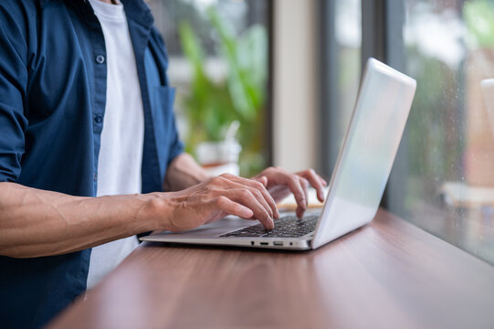 Close up of asian man's hands typing on laptop at wooden table while working in the cafe.