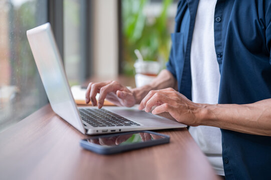 Close up of a freelancer man's hands typing on laptop beside a phone while at working at cafe table.