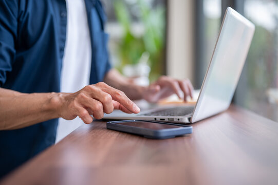Close up of asian man's hands working on laptop and touching a smartphone on wooden table in cafe