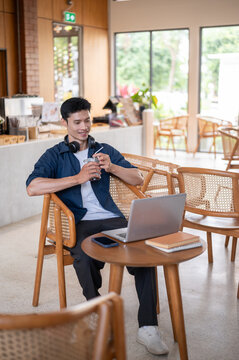 Asian man smiling and drinking iced coffee while watching online media on laptop at cafe table.