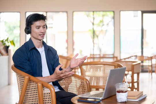 Asian freelancer man gesturing while talking on online meeting through laptop while working in cafe.