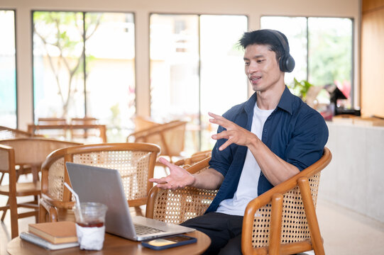 Side view of Asian man wearing headphones, talking on video conference via laptop in cafe.