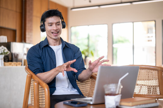 Asian man wearings headphones speaking happily during online meeting with laptop in modern cafe.