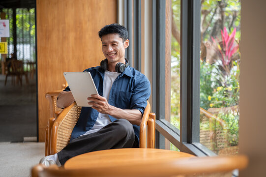 Asian man smiling while reading or looking at tablet while sitting leisurely at wooden table in cafe