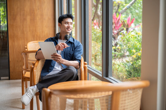 Asian man artist holding tablet and stylus pen looking over the glass wall while sitting in cafe.