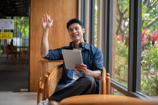 Asian man artist smiling while holding tablet and waving a hand while sitting at table in the cafe.