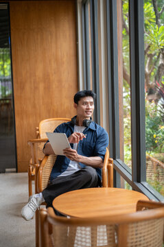 Asian freelancer man looking away from tablet while relaxing at wooden table in modern cafe.