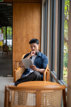 Asian freelancer man holding stylus looking thoughtfully at tablet while sitting at table in cafe.