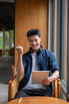 Excited Asian man making a fist celebrating success while looking at tablet sitting at table in cafe