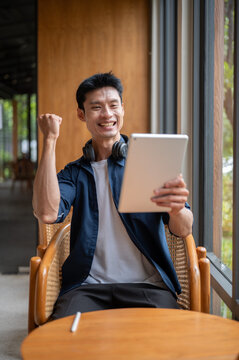Asian businessman celebrating achievement while looking happy at tablet and sitting at cafe table.