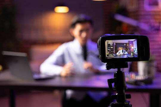 Male freelancer podcaster presenting online talk show from living room studio, young Asian man speaking into microphone and camera.