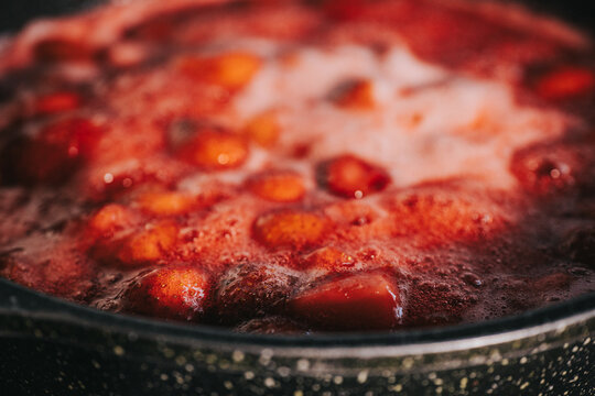 Close-up of whole strawberries simmering in bubbling red syrup with foam inside a metal pan, highlighting the jam-making process.
