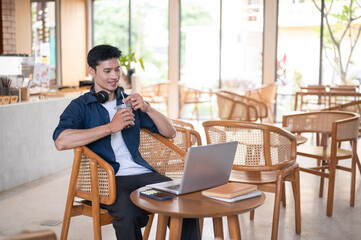 Fototapeta premium Asian freelancer man drinking coffee while watching or looking at laptop on wooden table in cafe.