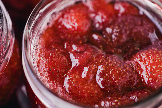 Close-up of whole strawberries immersed in glossy red syrup inside an open glass jar, with visible seeds, bubbles, and rich texture.