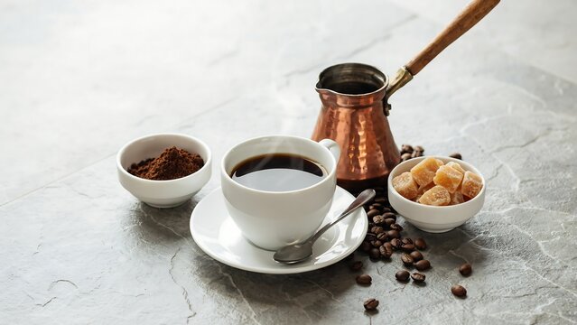 White coffee cup with spoon and copper pot beside bowls of coffee powder and sugar on grey stone