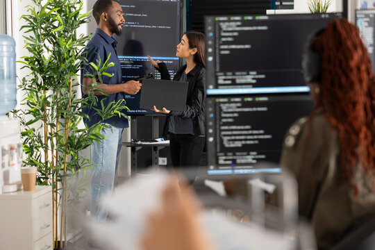 Male and female software developers stand by computer monitors, brainstorming coding project for startup. Diverse team collaborates on problem solving HTML scripts while building and refining website.