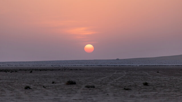 Beautiful pink and purple sunset dipping below a flat desert horizon, creating a serene, minimalist twilight landscape under pastel skies.