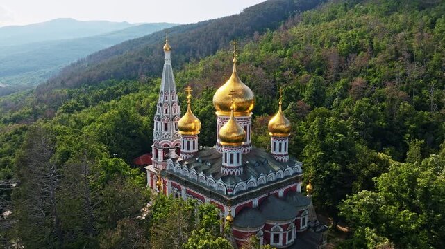 Golden domes of Shipka Memorial Temple of the Birth of Christ close up high angle aerial drone view lush green mountain forest Stara Planina background Bulgaria.
