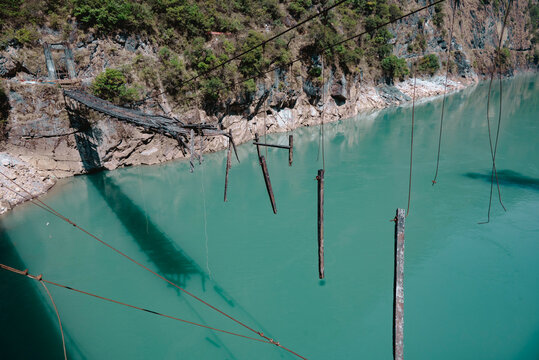 Broken wooden bridge over the turbulent Nujiang River in Yunnan, China, showing decayed planks and rusty cables against a dramatic mountain gorge landscape
