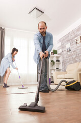 Obraz premium Husband uses a vacuum cleaner while cleaning apartment floor, showing domestic housework, floor hygiene and routine home cleaning responsibilities.
