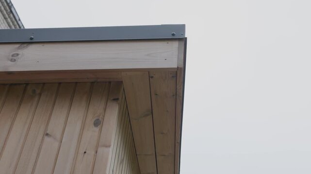 Low-angle close-up of a timber house roof eave with vertical wood cladding, exposed soffit boards, metal flashing, visible fasteners, and copy space in the overcast sky