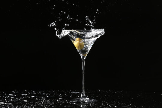 Glass of delicious martini with olive and splashes on dark background