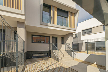 Side perspective of a contemporary house featuring a paved courtyard, wire mesh fences, and an elevated entrance accessed by stairs under a slatted balcony. © Toyakisfoto.photos