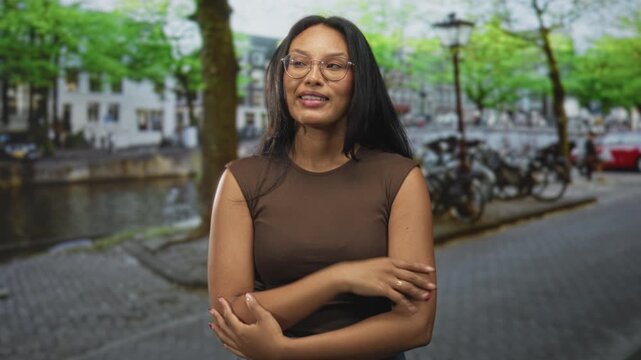 Woman wearing glasses crossing arms on street by canal with parked bicycles, lamp posts and cobblestone pavement; confidence reflection.