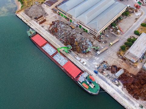 Cargo ship docks at port to unload materials near industrial site with large piles of scrap and storage buildings in the background during day
