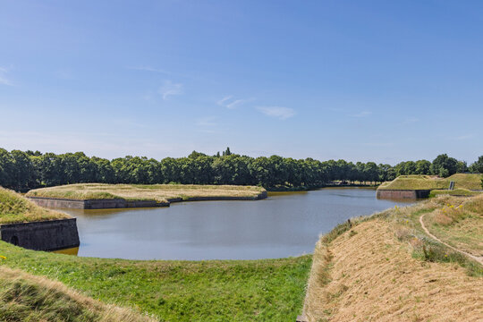 Naarden Vesting, Netherlands. Scenic view of the historical star fort fortifications, green grassy ramparts, and protective water moat under a clear blue sky.