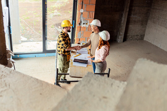 Smiling adult couple making agreement with handshake with a contractor at a house construction site.