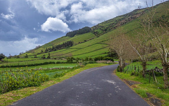 Camino de asfalto que serpentea a trav&eacute;s de un pintoresco paisaje rural de colinas verdes, pastos tradicionales y &aacute;rboles sin hojas bajo un cielo de tormenta en Azores.