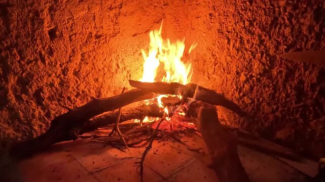 Close-up of bright orange and yellow flames burning thick wooden logs inside a rustic clay fireplace. Glowing embers and charred wood illuminate rough earthen walls with warm firelight.