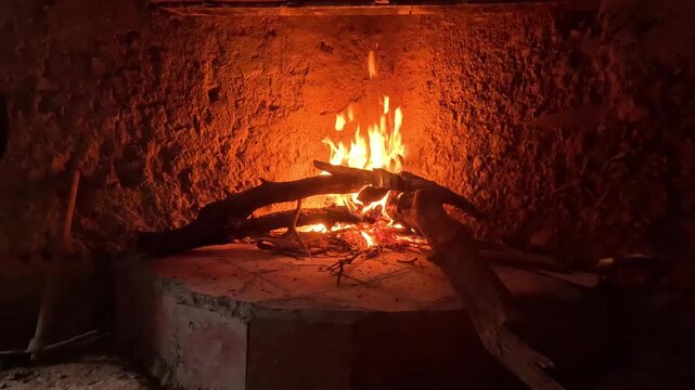 Close-up of bright orange and yellow flames burning thick wooden logs inside a rustic clay fireplace. Glowing embers and charred wood illuminate rough earthen walls with warm firelight.