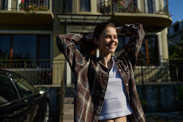 Fototapeta premium Casual outdoor portrait of a smiling woman wearing a plaid shirt, enjoying daytime sunlight, relaxed pose in front of a residential building, lifestyle mood