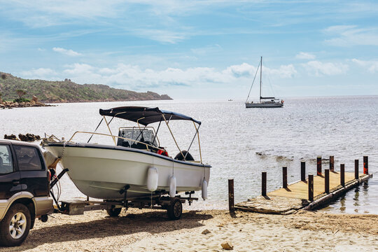 Motorboat on a trailer towed by an off-road vehicle at a sandy beach launch with a sailboat offshore. Boat launching and coastal recreation for the marine leisure and watersports market