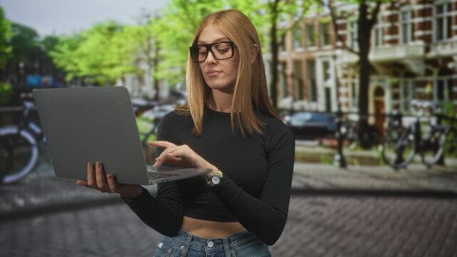 Woman holding laptop in left hand and showing thumbs down gesture on cobbled city street, wearing glasses and a black crop top; confidence independence.