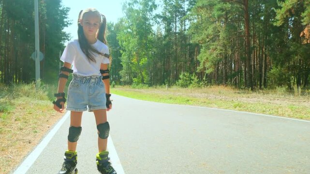 Child girl in casual clothes roller skating on road in forest on sunny summer day.