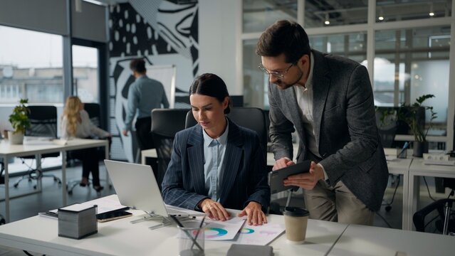Two Caucasian business colleagues talking discuss data in digital tablet compare information in laptop businesswoman female with businessman male leader talk together analysing online work in office