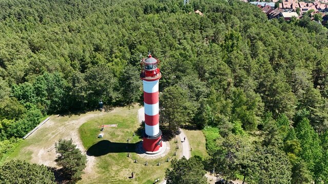 Aerial high angle view of Nida lighthouse with a long shadow in a green pine forest on Curonian Spit, Lithuania. Scenic summer landscape with a historic red and white beacon.