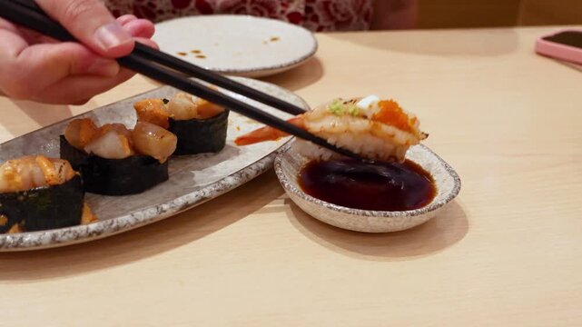 Dipping fresh prawn sushi into savory soy sauce with hotate sushi plate in background