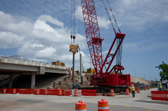Red crawler crane lifting piles as construction workers perform bridge repair and roadwork beside a highway. Editorial Use Only July 15, 2026 St. Petersburg, FL  USA under a bright partly cloudy sky.
