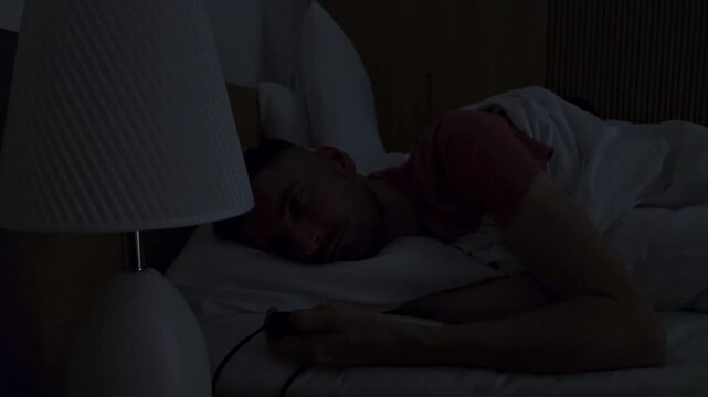 Depressed man lying awake in bed at night repeatedly switching bedside table lamp on and off while staring into space. Young adult struggling with insomnia, anxiety, emotional burnout, hopelessness