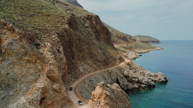 Horizontal aerial view of two small rental cars driving along a precarious narrow dirt track cut into a steep rocky coastal cliff edge on the path to Balos bay