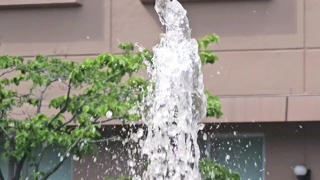 Close up of water fountain jet splashing in urban park with trees and building in background. Slow motion style footage capturing flowing water, droplets and refreshing city environment