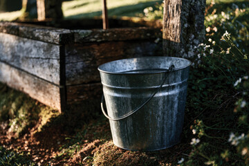 Fototapeta premium A weathered bucket sits beside an ancient wooden well surrounded by lush greenery, capturing the essence of rustic charm and connection to nature and its resources.