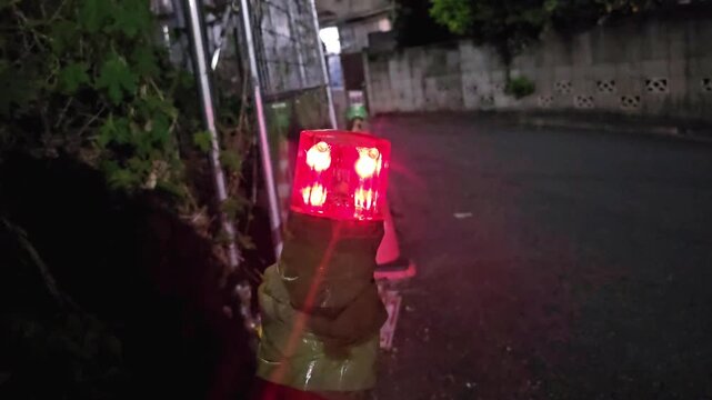 Flashing red LED safety beacon attached to roadside barrier during nighttime road construction with traffic cone and street in background. Temporary hazard warning, traffic control