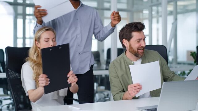 Three multiracial business team dancing with papers in office Caucasian and African American colleagues coworkers multiethnic group teamwork job success dance holding documents having fun partnership