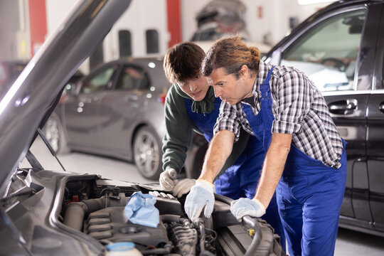 Experienced middle-aged automotive mechanic mentoring young apprentice. Men conversing while examining car engine under open hood in well-lit auto repair workshop