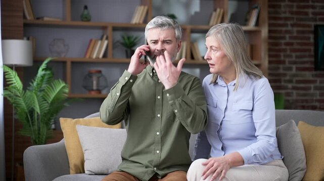Angry senior man talking on a smartphone while sitting on a sofa beside his wife in the living room at home. Dissatisfied retired couple complaining about poor service and leaving a negative review.
